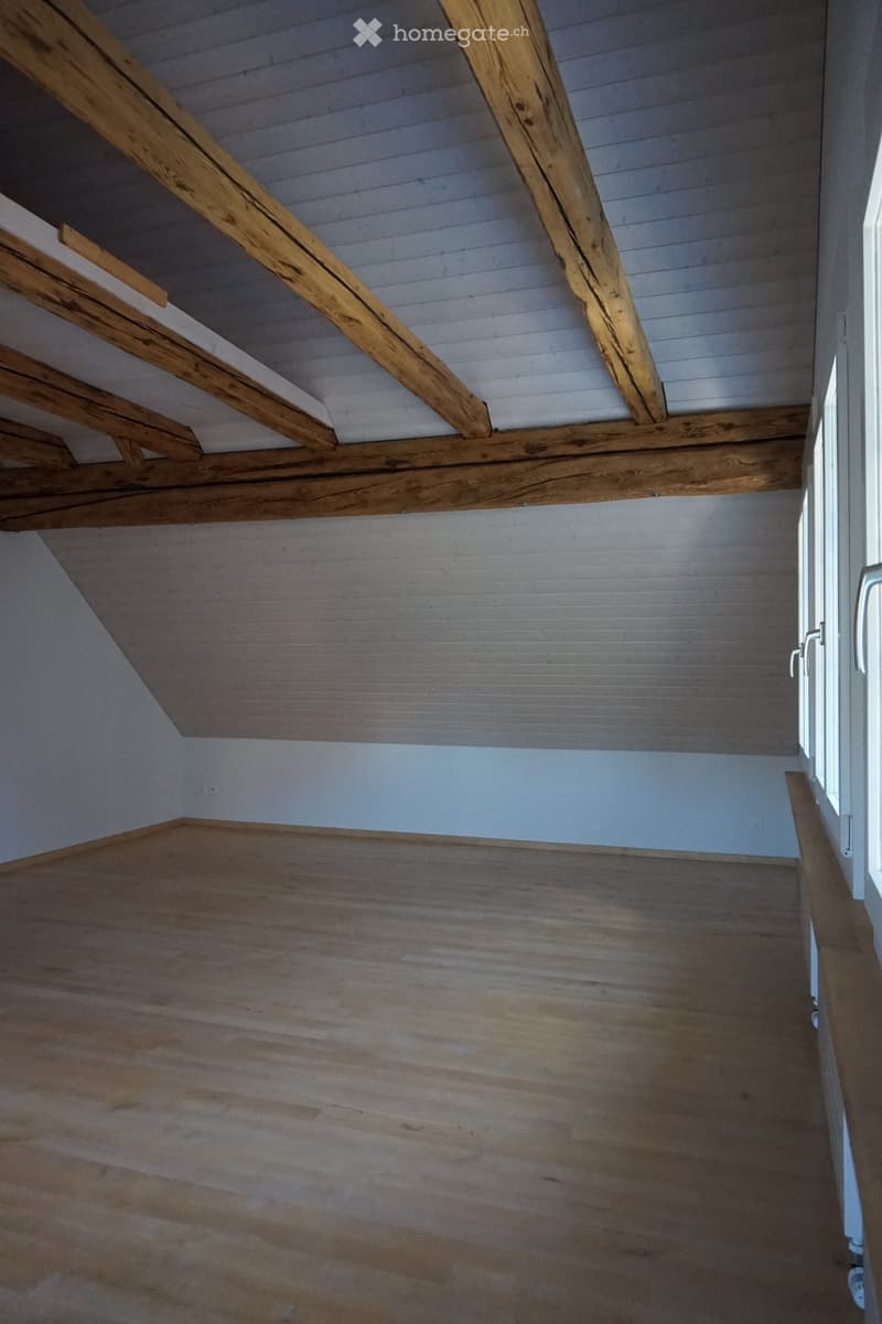 Empty room with wooden floor and exposed wooden beams on the ceiling. Light blue walls and a window on the right side.