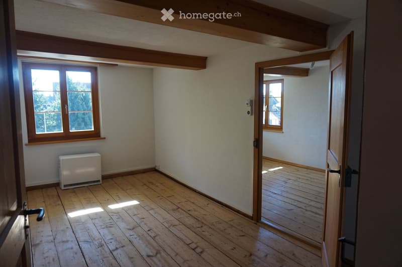 Empty room with wooden flooring, exposed wooden beams on the ceiling, and a radiator on the wall. There are two windows and an open doorway leading to another room.