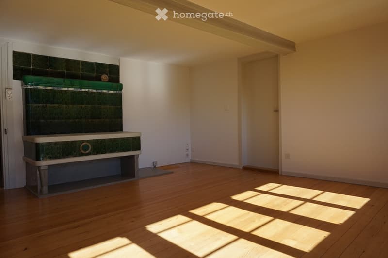 Empty room with wooden flooring, white walls, and a decorative fireplace with green tiles and a white mantel. Sunlight streams in from a window, casting shadows on the floor.