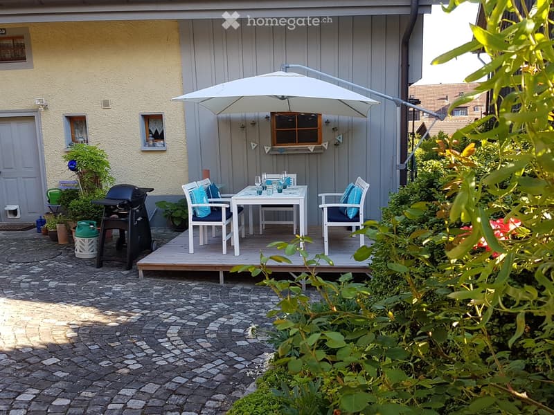 Outdoor patio area with a table, chairs, umbrella, and grill, surrounded by greenery and a paved stone pathway.