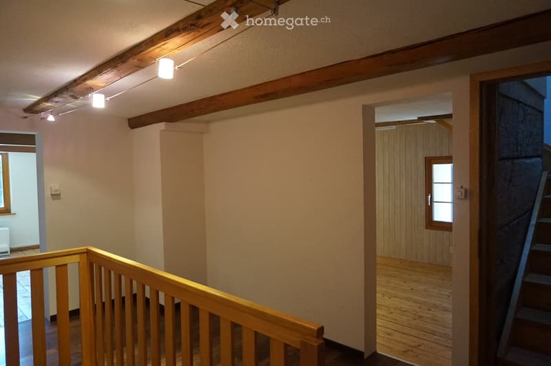 Wooden staircase with a handrail, exposed wooden beams on the ceiling, and warm lighting. The hallway has light-colored walls and leads to another room with wooden paneling.
