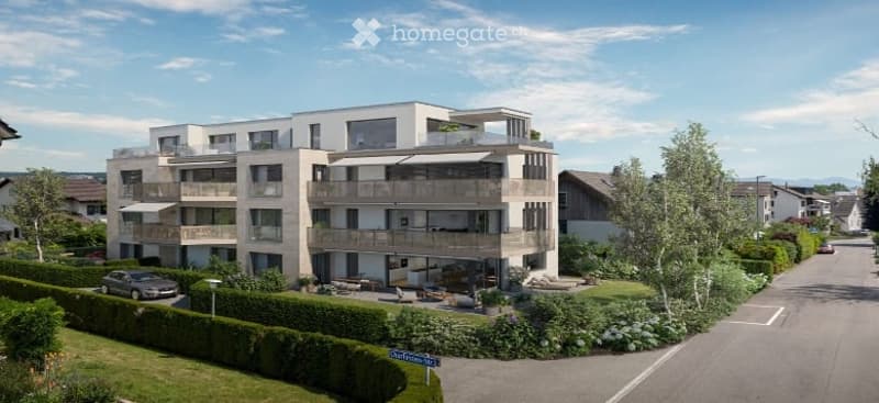 Modern multi-story residential building with balconies, surrounded by greenery and a paved driveway.