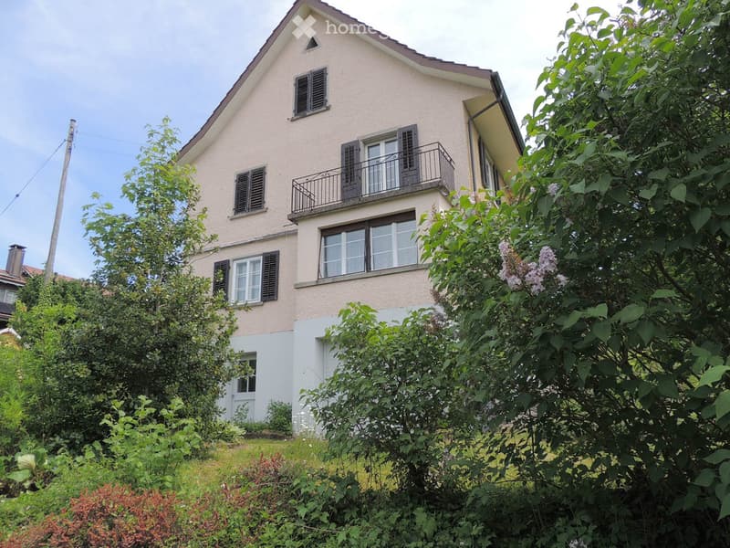 Two-story house with beige siding, multiple windows with shutters, and a small balcony on the upper floor. The house is surrounded by lush greenery and trees.