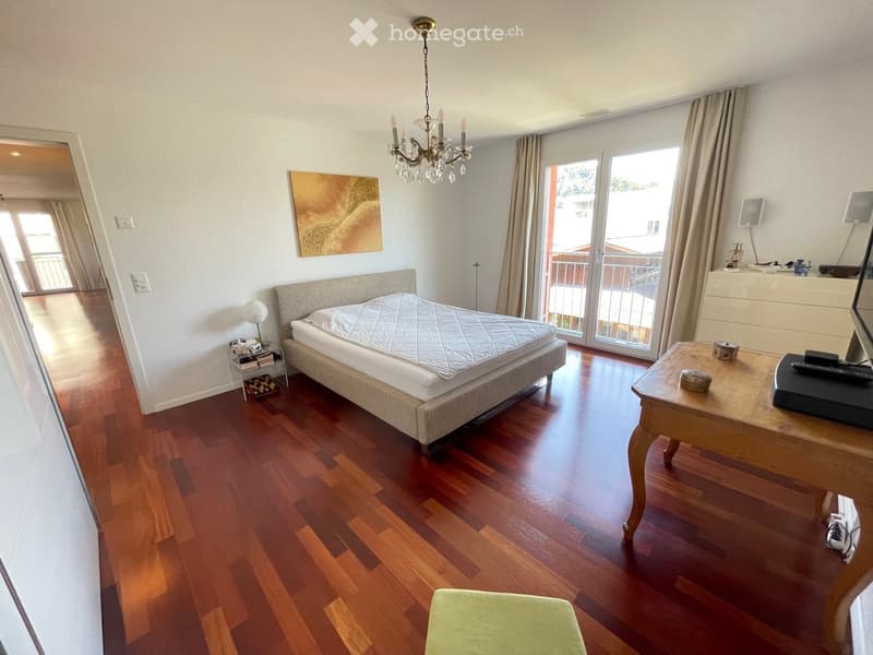 Spacious bedroom with wooden flooring, large bed, side table, desk, and balcony access.