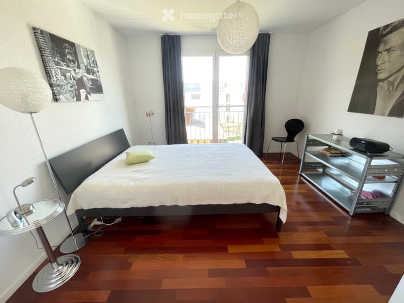 Modern bedroom with wooden flooring, a large bed with white bedding, a round glass stool, a sleek chair, a small table with a radio, and two large picture frames on the walls.