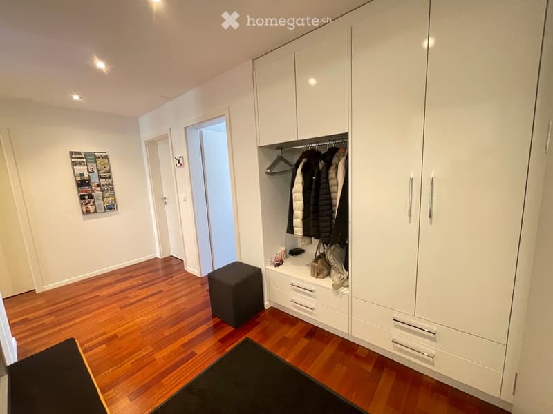 A modern hallway with wooden flooring, white walls, a built-in white cabinet with coat rack, a small black stool, and a picture frame on the wall.