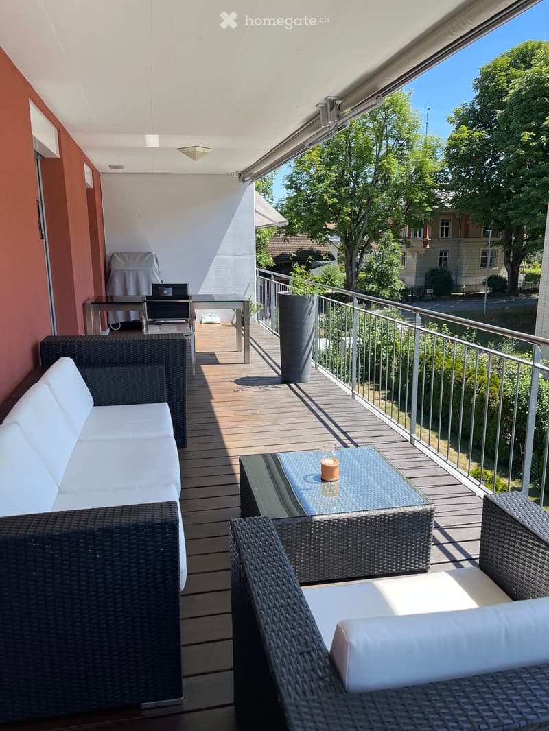 Balcony with wooden decking, white sofa, glass coffee table, wicker chairs, and a covered grill area.
