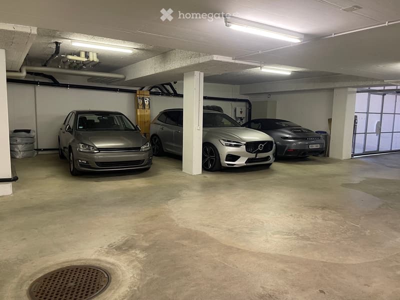 Underground parking area with four parked cars, concrete floor, white walls, and fluorescent lighting.