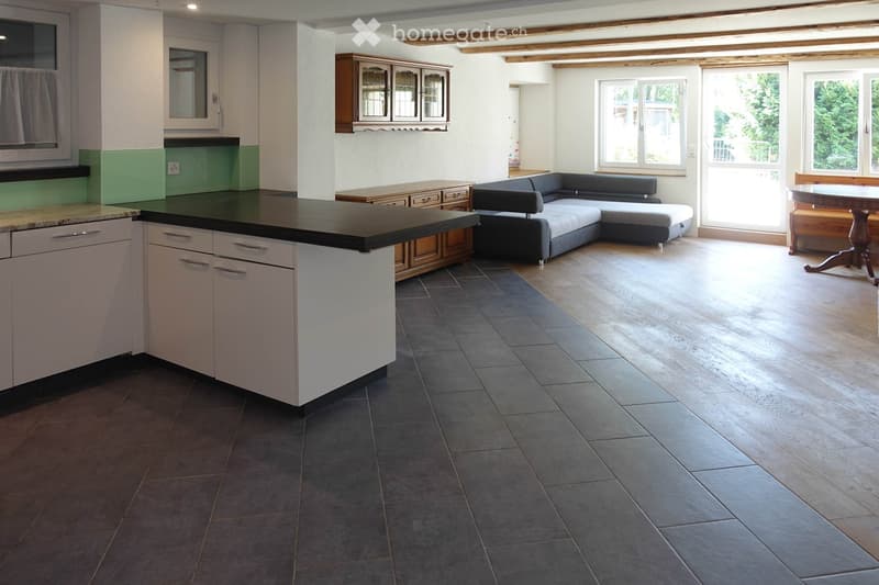 Open-plan living and dining area with a grey sectional sofa, coffee table, kitchen island, and dining table with chairs. The space features a mix of tiled and wooden flooring, large windows, and built-in cabinets.