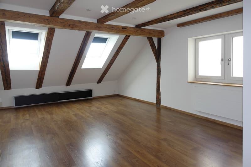 Empty attic room with wooden beams, wooden flooring, white walls, and skylight windows.