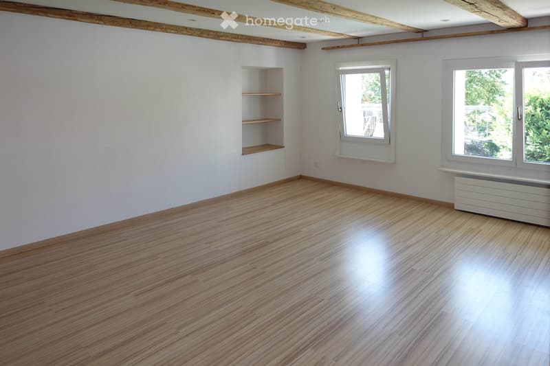 Empty room with wooden flooring, white walls, exposed wooden beams on the ceiling, and built-in shelving unit. Two windows with white frames and a radiator under one window.