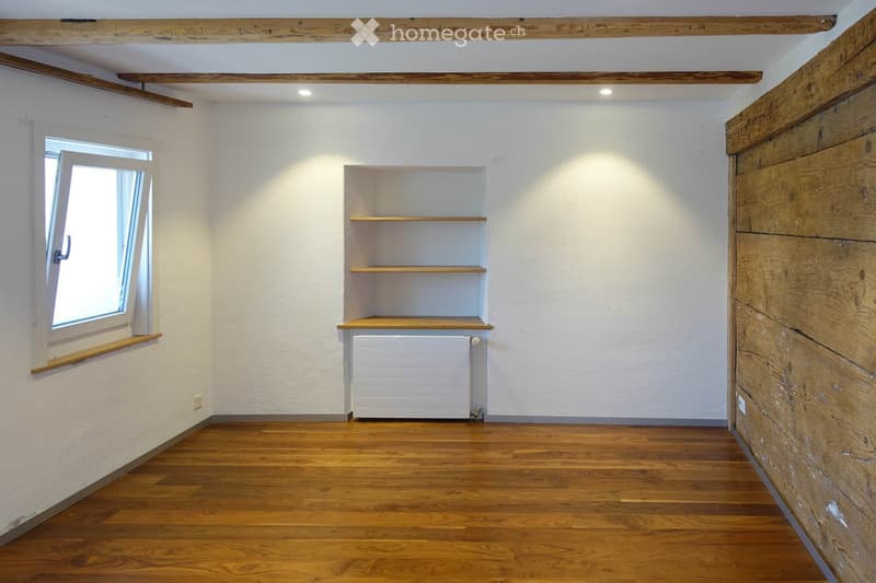 Empty room with wooden flooring, white walls, exposed wooden beams on the ceiling, a window with wooden sill, and a built-in wooden shelf unit with a radiator below.