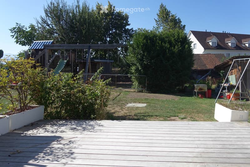 A wooden deck in the foreground leads to a grassy yard with a playground area, including a swing set and slide, surrounded by trees and a house in the background.