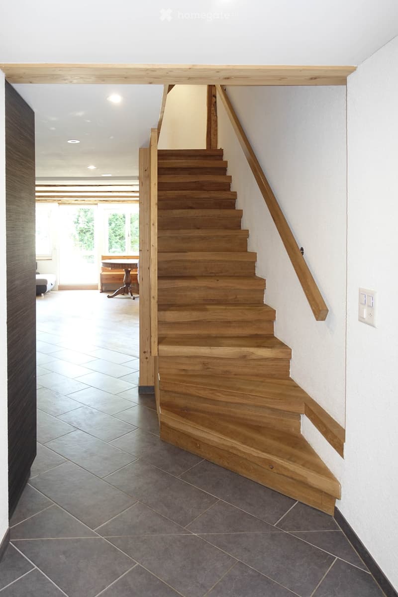 Wooden staircase with a handrail leading to an open area with a dining table and chairs in the background.