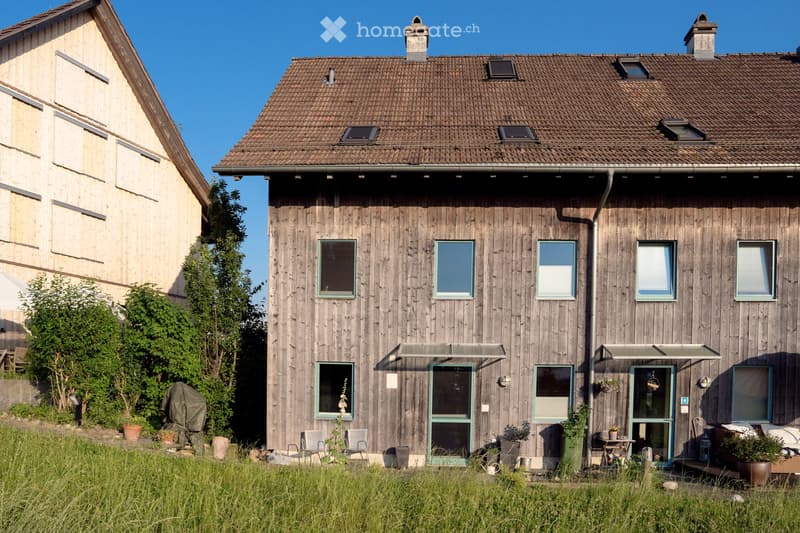 Two-story wooden house with a tiled roof, multiple windows, and a small porch area with outdoor furniture and potted plants.