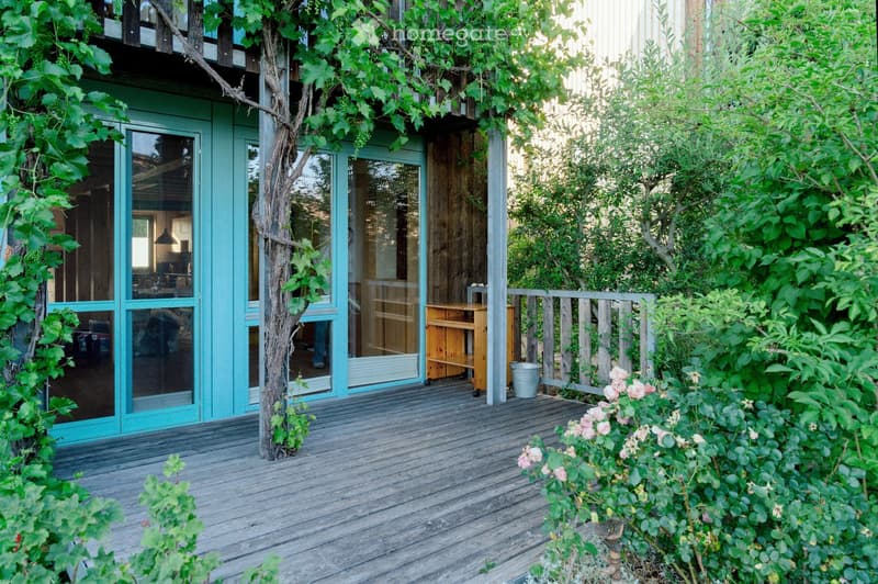 Wooden deck with a small table and chair, surrounded by lush greenery and climbing vines, with a view of the house interior through large glass doors.