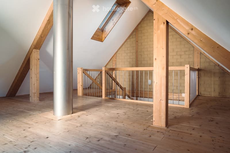An empty attic space with wooden floors, exposed wooden beams, and a metal staircase leading to another level. The room features a skylight and a central cylindrical column.