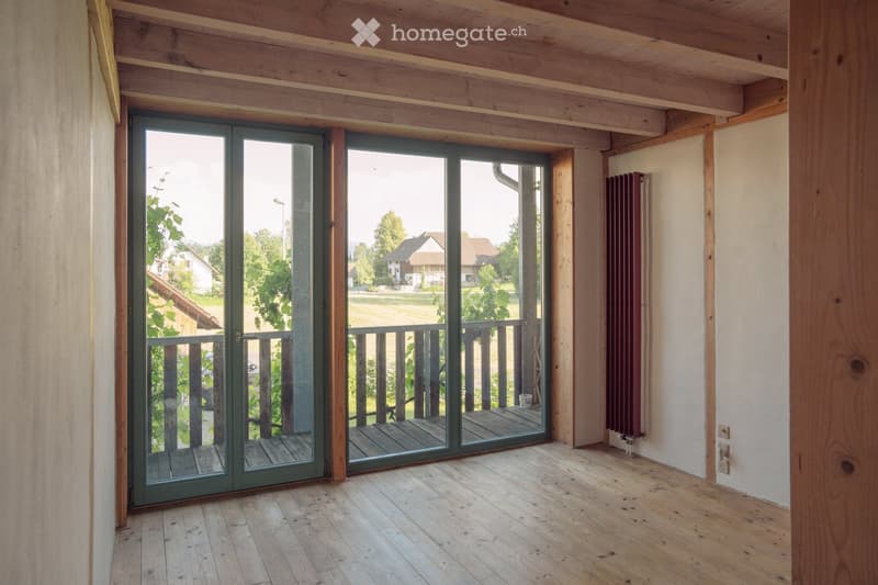 Empty room with wooden flooring, exposed wooden beams on the ceiling, large glass sliding doors leading to a wooden deck, radiator on the wall, and a view of a green outdoor area.