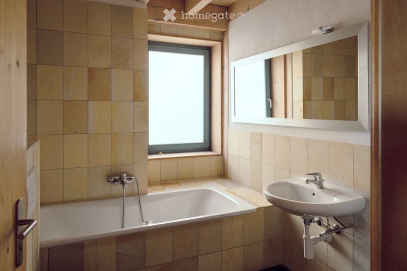 Bathroom with beige tiled walls and floor, a bathtub, a sink with a faucet, and a large mirror above the sink. There is a window providing natural light.
