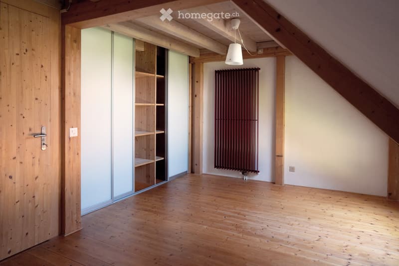 Empty attic room with wooden floors, exposed wooden beams, a sliding glass door leading to a closet, and a radiator on the wall.