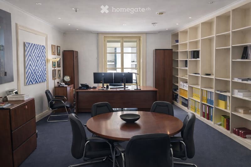 Office space with a large wooden desk, two computer monitors, a round meeting table with chairs, shelves filled with binders and books, filing cabinets, and artwork on the walls.