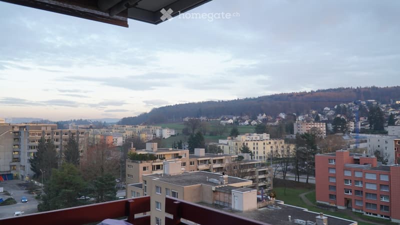 View from a balcony overlooking a residential area with apartment buildings, trees, and distant hills.