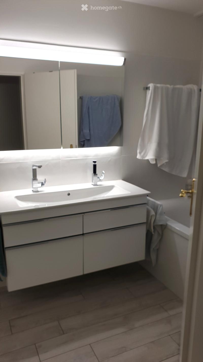 Modern bathroom with a white sink, two faucets, a large mirror with built-in lighting, and a towel rack with towels.