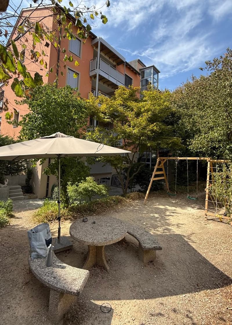 Garden area with a stone table, bench, umbrella, swing set, and surrounding greenery.