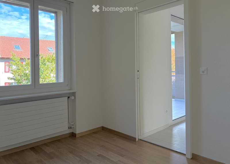 Empty room with wooden flooring, white walls, large window with view of neighboring building, radiator, and sliding glass door leading to balcony.
