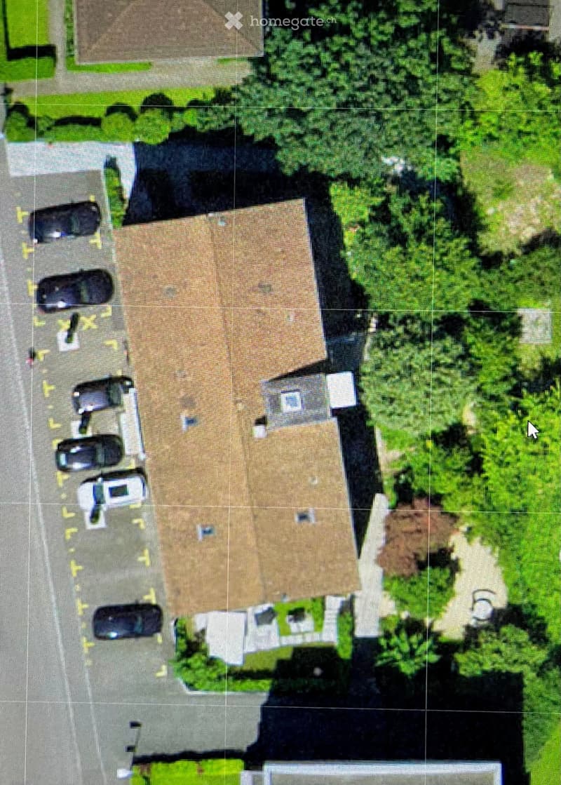 Aerial view of a two-story house with a brown roof, surrounded by greenery and parked cars.