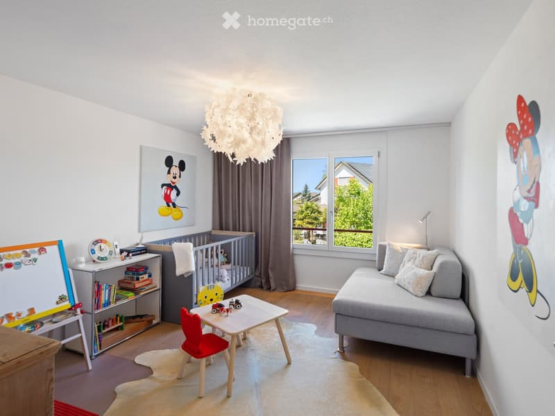 Brightly lit children's room with a grey sofa, small table, red chair, bookshelf, crib, and artwork featuring Mickey Mouse.