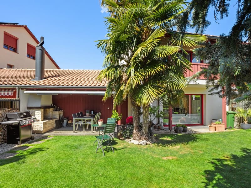 Well-maintained garden with a green lawn, a large palm tree, a dining set with a table and chairs, a grill, and several potted plants.