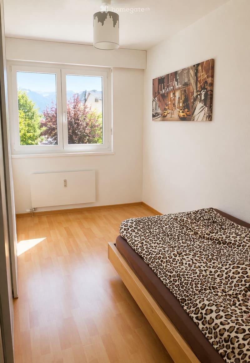 Bright bedroom with wooden flooring, large windows showing a scenic view, a bed with a leopard print cover, and a large picture frame on the wall.