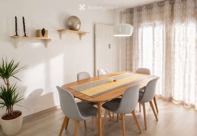 Dining room with a wooden dining table, six grey chairs, a yellow and grey table runner, a potted plant, and decorative items on a shelf.