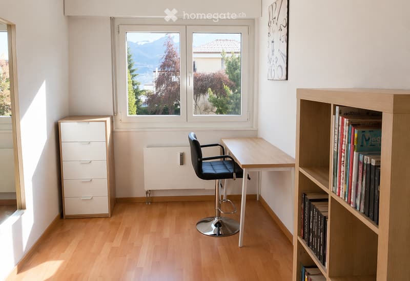 Bright study room with wooden flooring, large windows offering a scenic mountain view, a desk with a chair, a bookcase filled with books, and a drawer unit.
