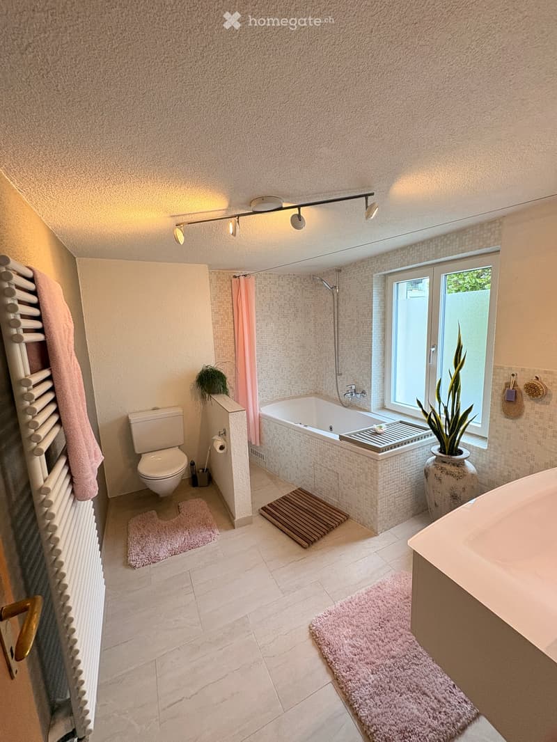 Modern bathroom with a bathtub, toilet, radiator, and a window with a plant. Features include a pink towel, a pink rug, and a decorative plant in a vase.