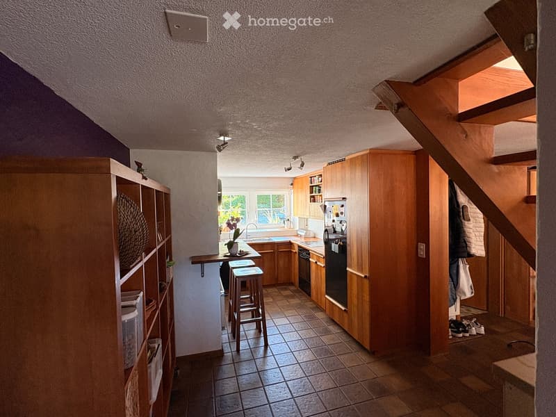 Modern kitchen with wooden cabinets, stainless steel appliances, and a bar area with stools. Features a large window, a small table with a vase, and a staircase leading to another level.