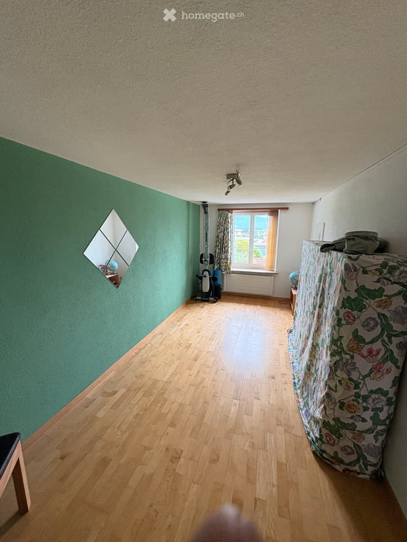 A furnished room with wooden flooring, green wall, and a window with curtains. Contains a stool, mirror, globe, radiator, and a covered mattress.