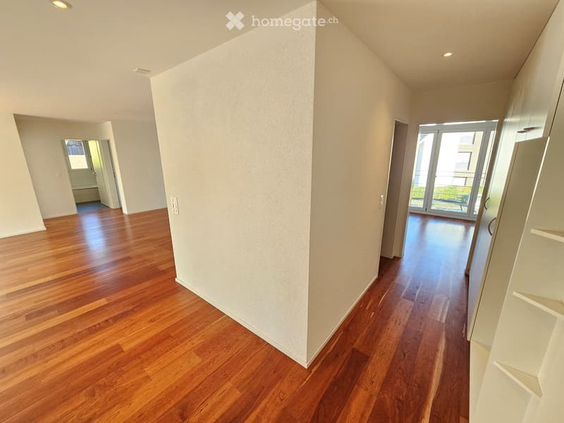 Empty hallway with wooden flooring, white walls, and recessed lighting. Features a sliding glass door leading to an outdoor area and an open doorway to another room.