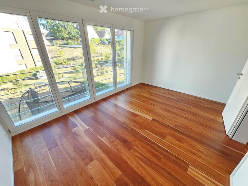 Empty room with wooden flooring and large glass doors leading to a balcony or terrace. The space is unfurnished, with white walls and a view of the outdoor area.