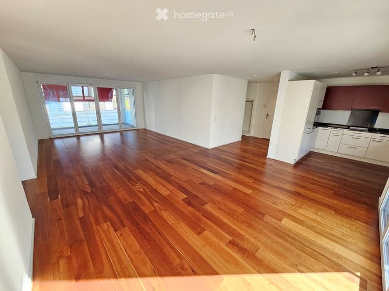 Open-plan living and dining area with wooden flooring, white walls, and large sliding glass doors leading to a balcony. Kitchen area with white cabinets and stainless steel appliances is visible on the right side.