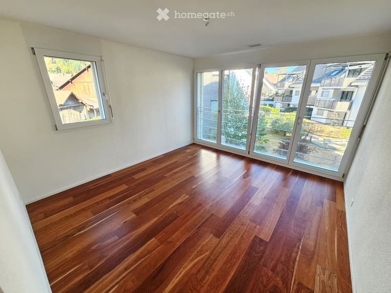 Empty room with wooden flooring and large windows, including a sliding glass door leading to a balcony.