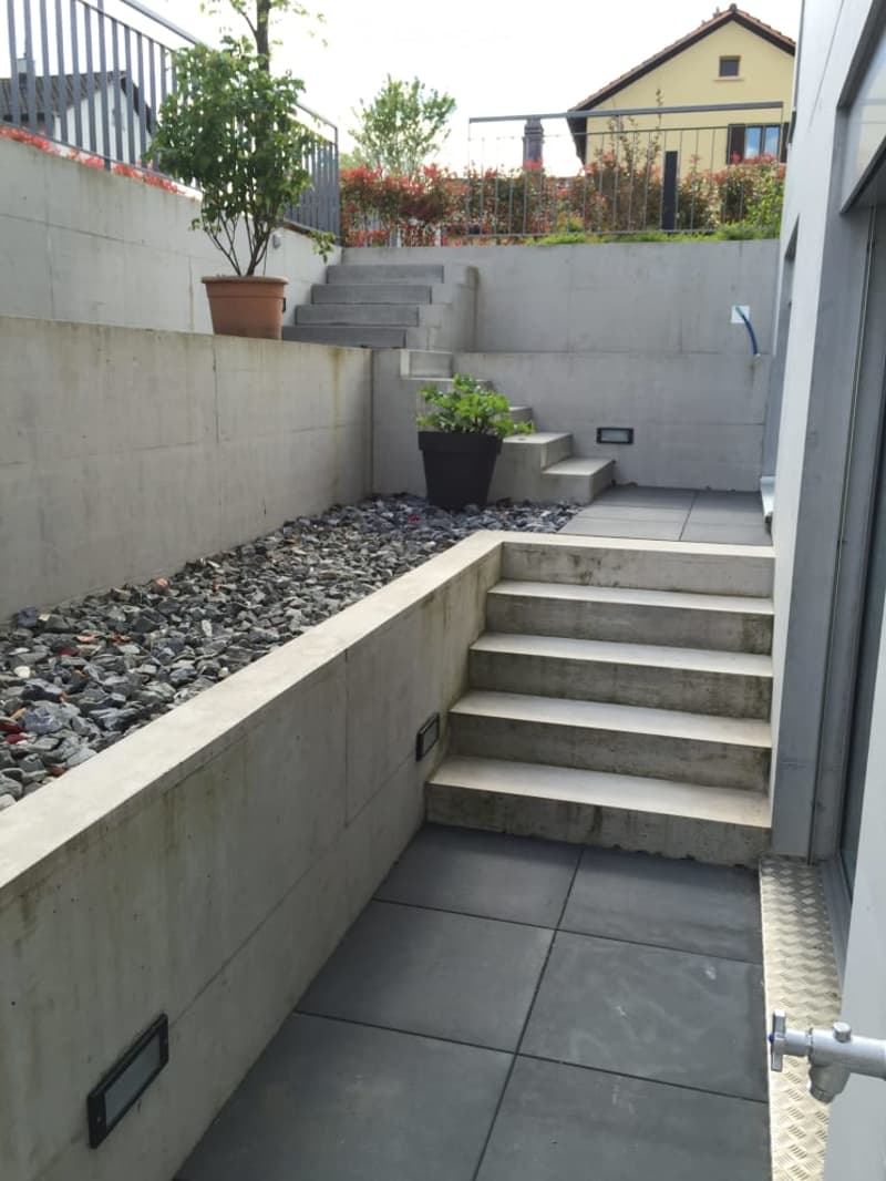Modern concrete patio with steps, potted plants, and a raised garden bed bordered by gravel.
