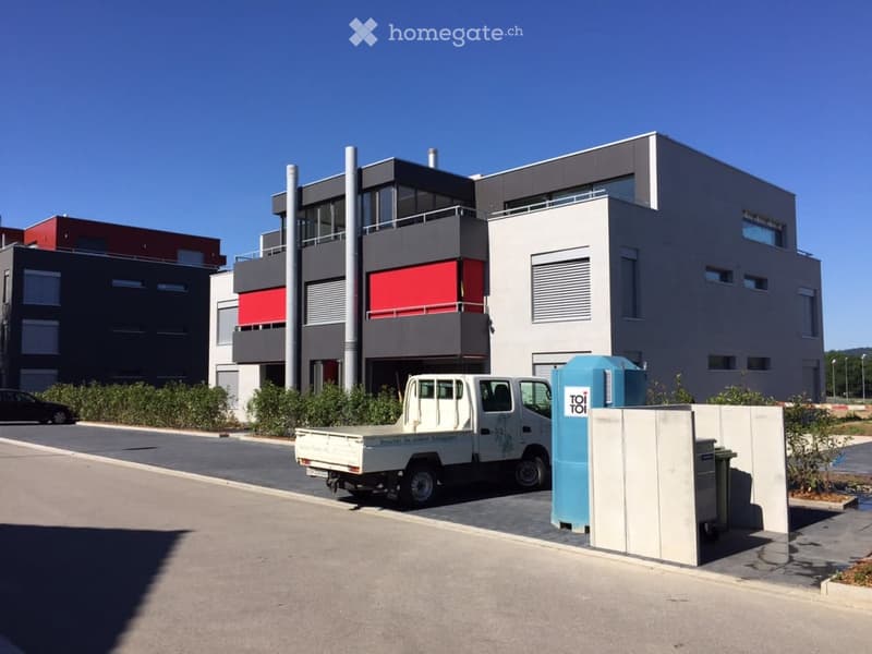 Modern multi-story building with a combination of grey and red panels, large windows, and a balcony on the upper floor. A white truck is parked in front, and there are portable toilets and a trash bin nearby.