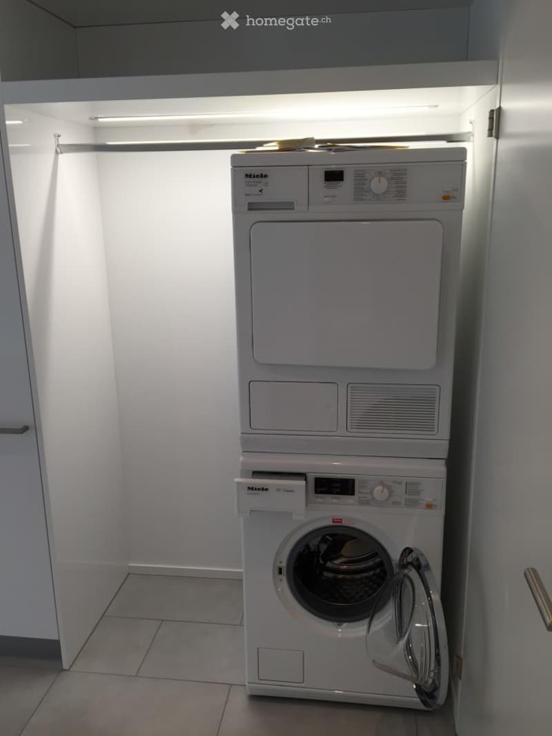 Modern bathroom with a white washing machine and dryer unit, both with control panels and digital displays, placed in a well-lit, tiled space.