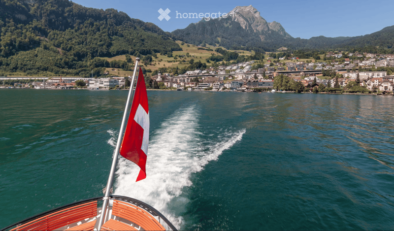 A scenic view of a town on the shore of a lake, with mountains in the background and a Swiss flag on a boat in the foreground.