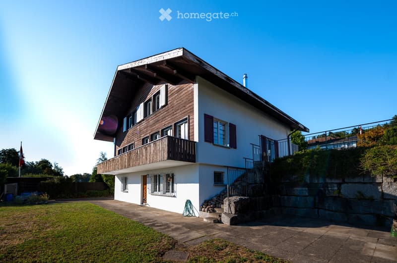 Two-story house with white walls, wooden accents, multiple windows, and a balcony on the upper level. The house is surrounded by a well-maintained lawn and stone steps leading up to the entrance.