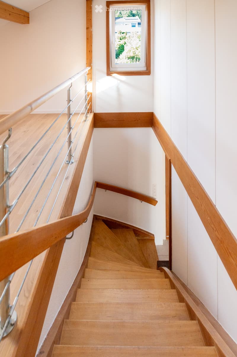 Wooden staircase with metal railing, light-colored walls, and a window showing greenery outside.