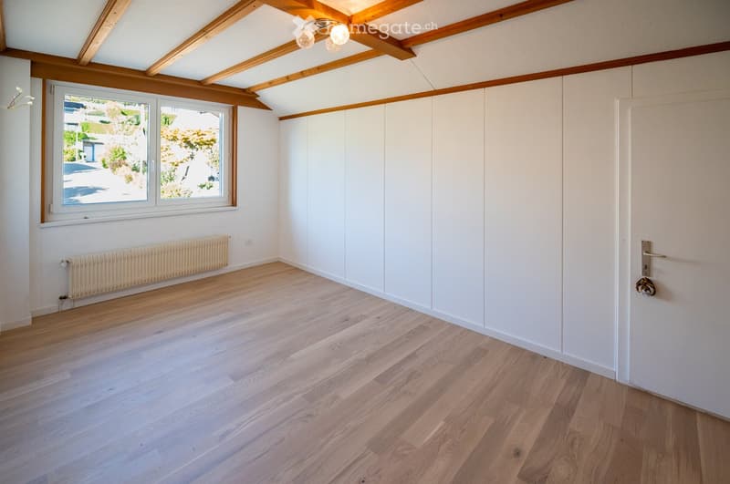 Empty room with light wooden flooring, white walls, built-in white closet, large window with view, and exposed wooden ceiling beams.