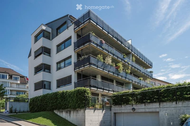 Modern multi-story apartment building with balconies, large windows, and greenery on the lower level.